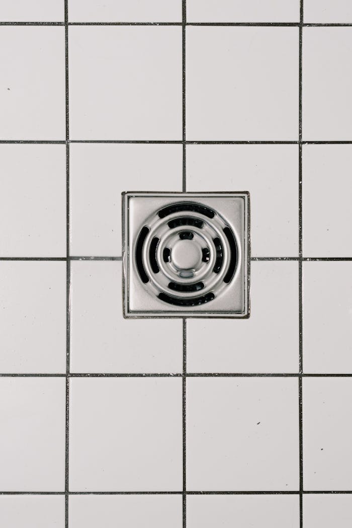 our-story-01 Top view of white ceramic tile floor with stainless steel square shaped cover of drain in contemporary shower
